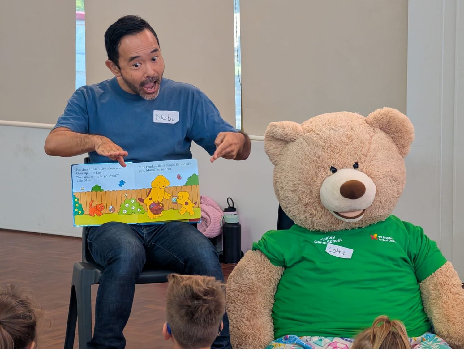A seated instructor named uses expressive hand signs and facial gestures while reading a children's book to a group, sitting next to a large teddy bear mascot wearing a green shirt named Cottie.